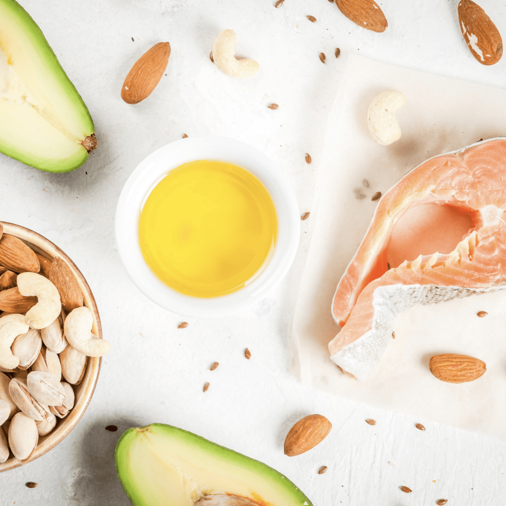 Raw salmon next to a small bowl of oil and sliced avocados, surrounded by nuts on a white background.