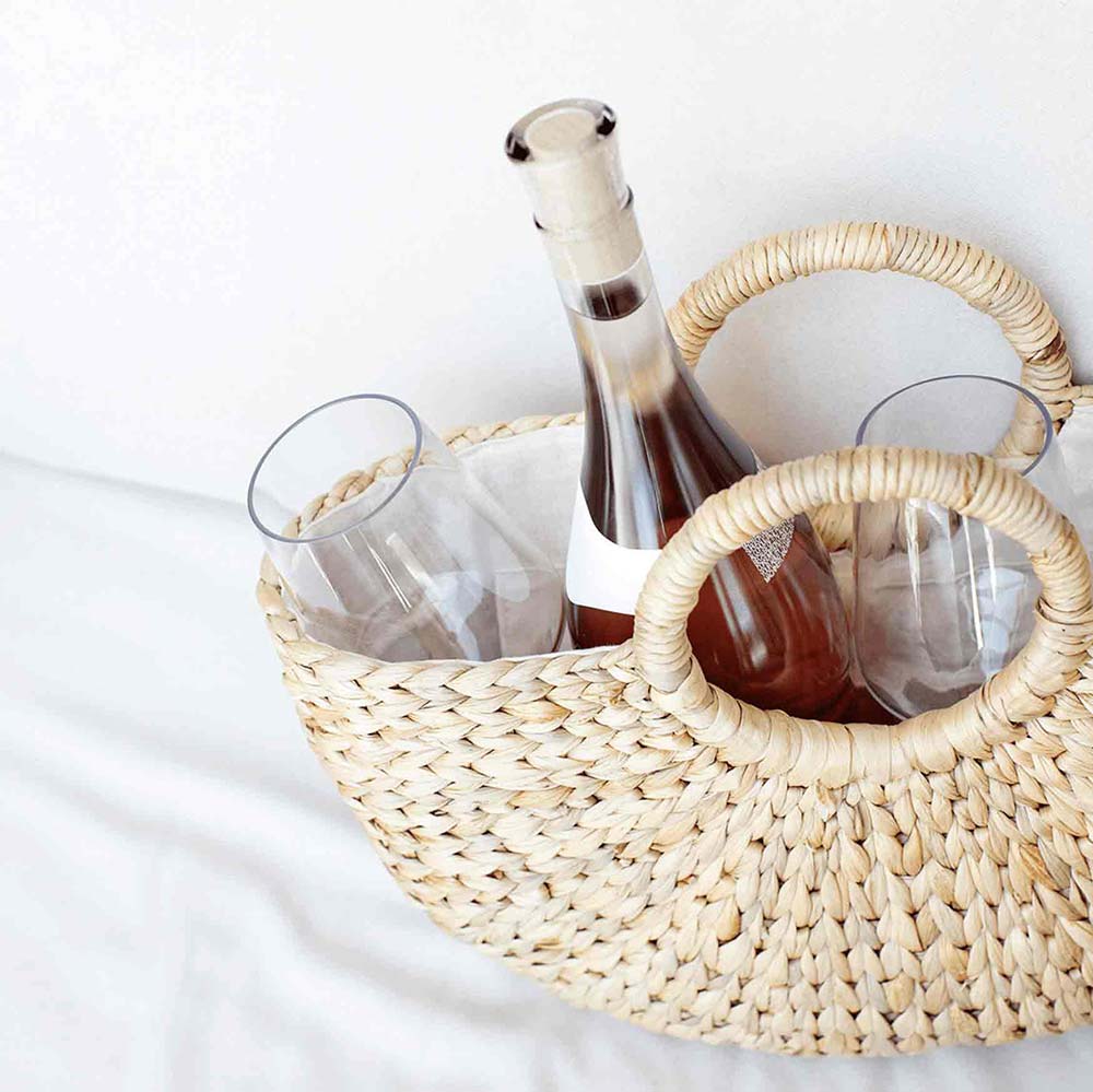 A tan basket filled with a bottle of wine and two wine glasses on a white cloth and white background.