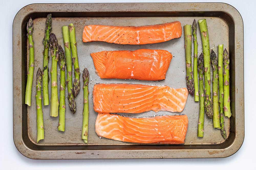 Four pieces of raw salmon in the middle of a sheet pan in the middle of asparagus.