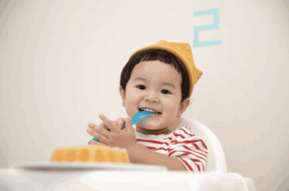 A child with black hair and a gold crown wearing a red and white stripped shirt eats yellow cake with a blue spoon.
