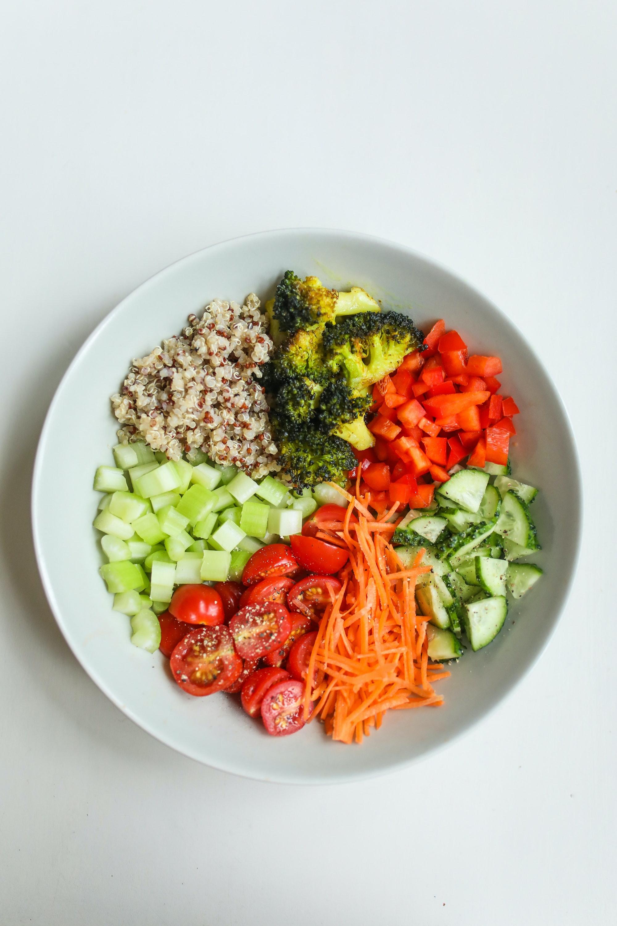 White bowl of carrots, cucumber, tomatoes, red bell pepper, broccoli, and quinoa on a white background.