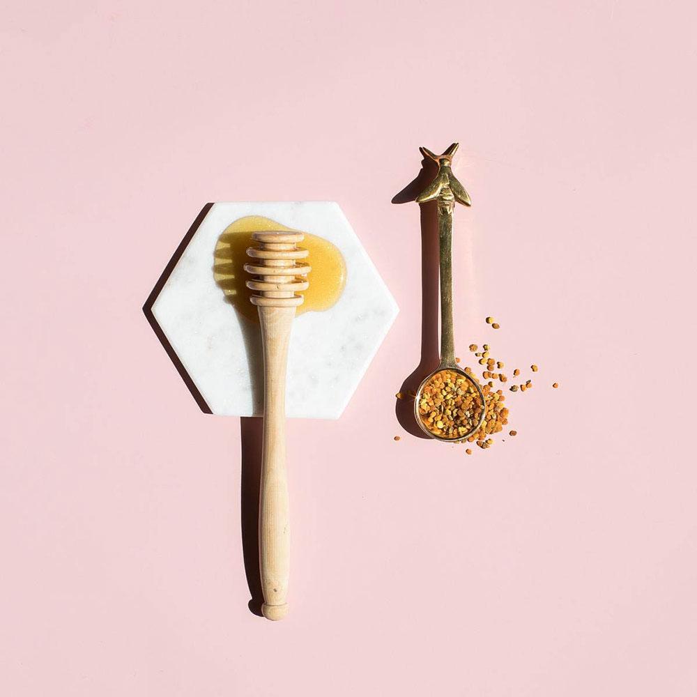 Honey on a white marble coaster next to spoon of orange seeds on a pink background.