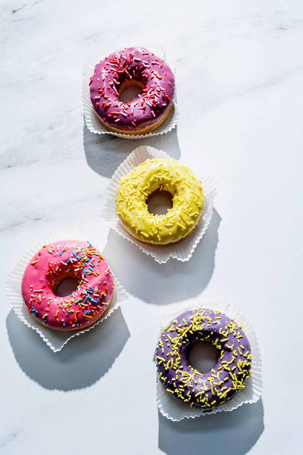 4 donuts, one has yellow frosting, purple frosting, pink frosting and dark pink frosting on a white background.