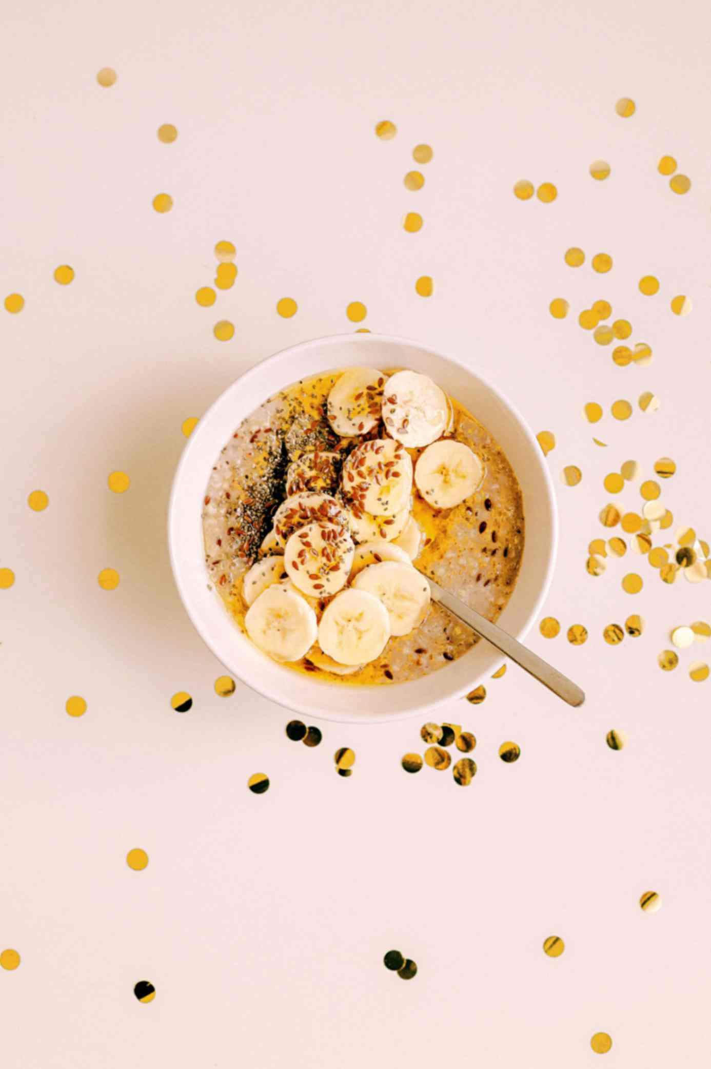 A bowl of outmeal with banana slices and seeds on top. Gold confetti is on the table surrounding the bowl.