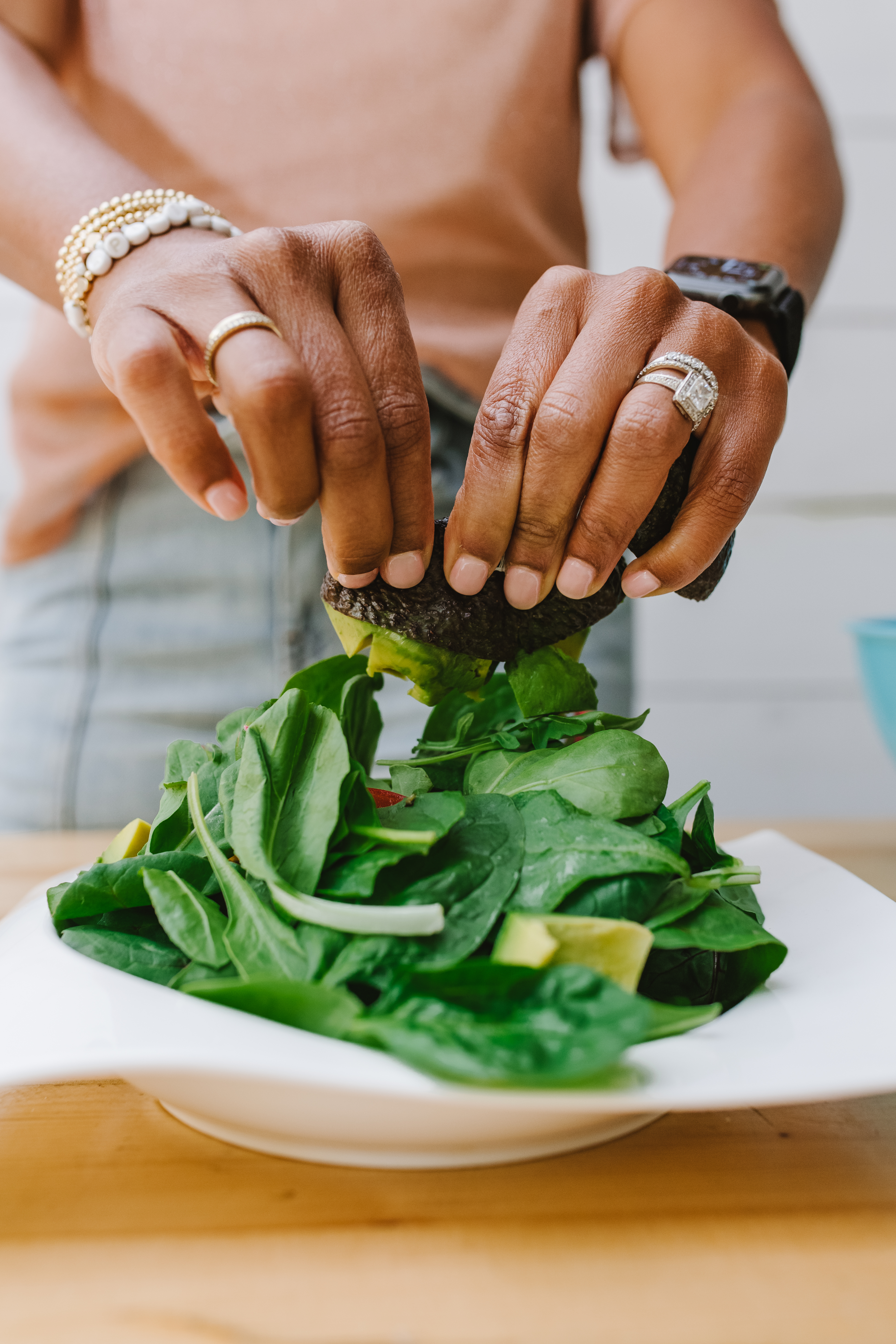 Woman wearing rings squeezing avocado on to a green salad on a white plate, on a wood counter.
