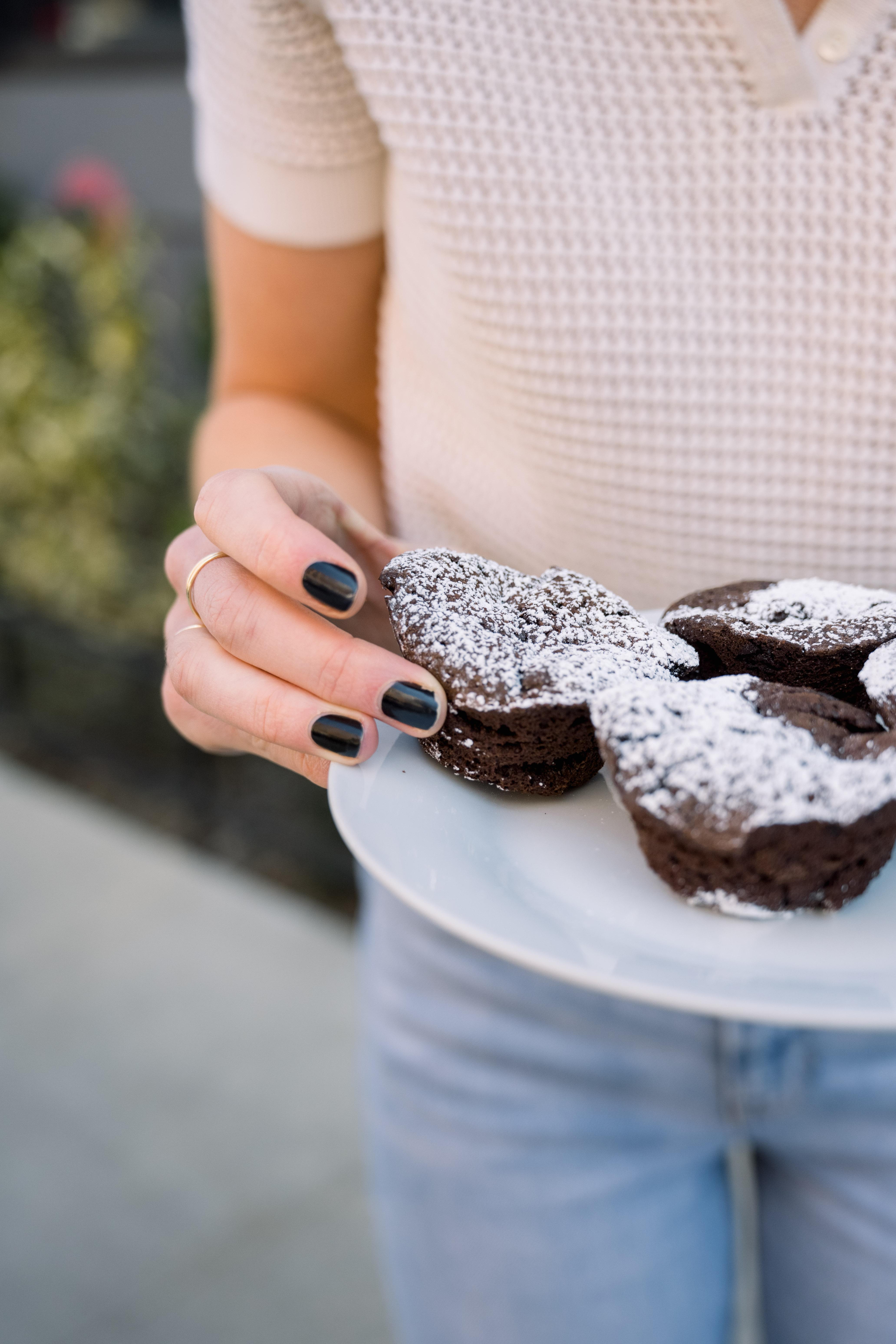 A person holding a plate of chocolate brownie cupcakes with powdered sugar on top.