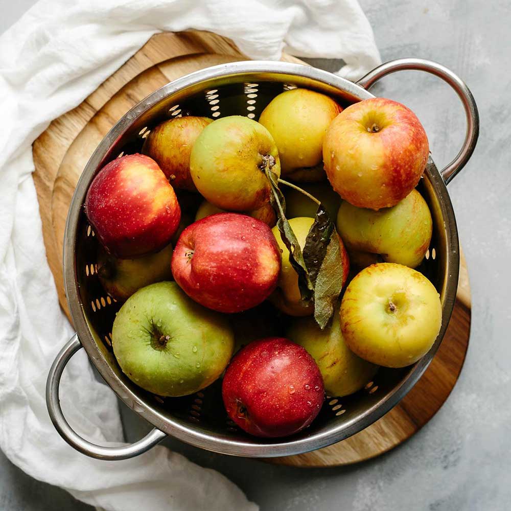 A silver strainer with washed apples inside on a wood cutting board surrounded by white cloth.