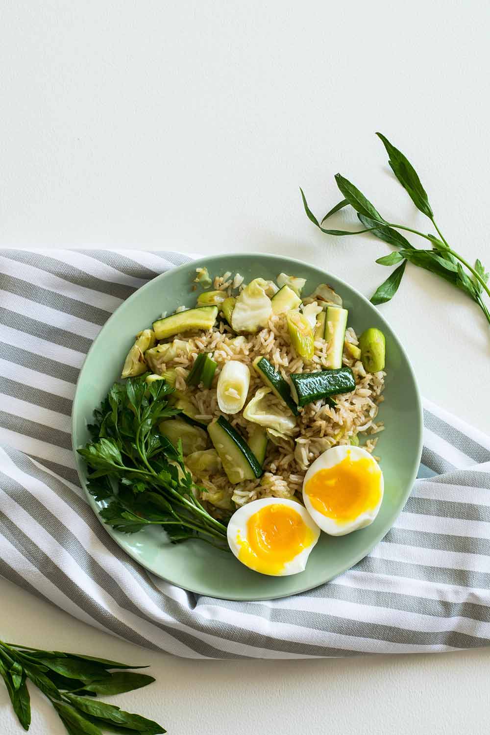 Green plate filled with foods like rice, zucchini, greens and an egg on top of a striped kitchen towel with greenery around it.