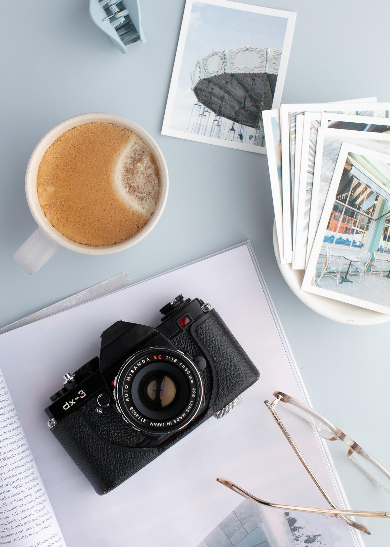 White notebook with black camera on top next to mug with coffee surrounded by different printed pictures.