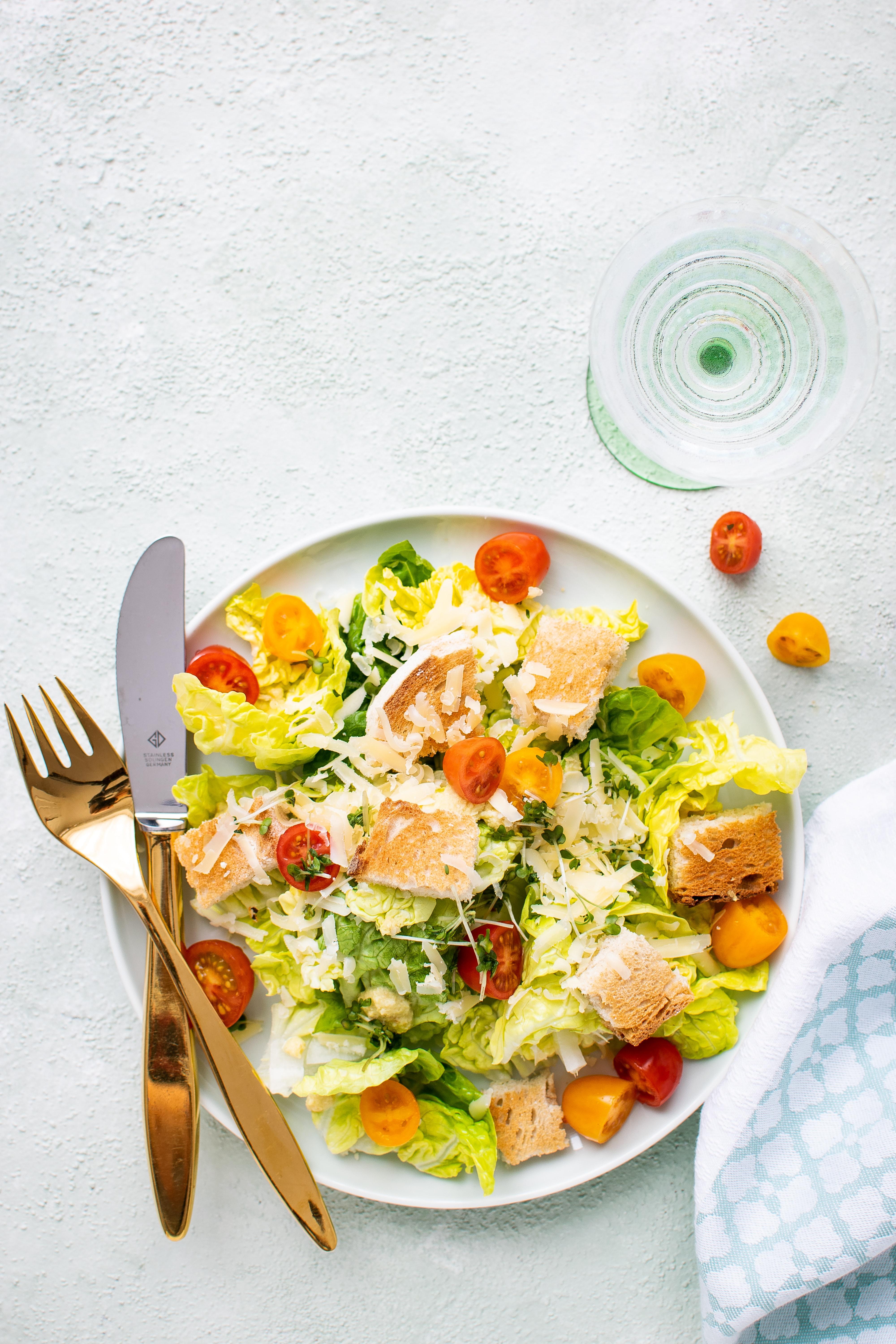 White plate with a mixed greens salad on top with gold fork and knife sitting on top next to a glass of water on a white background.