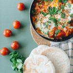Shakshuka in a saute pan with a side of pita bread. Cherry tomatoes and a bunch of parsley are also scattered on the table.