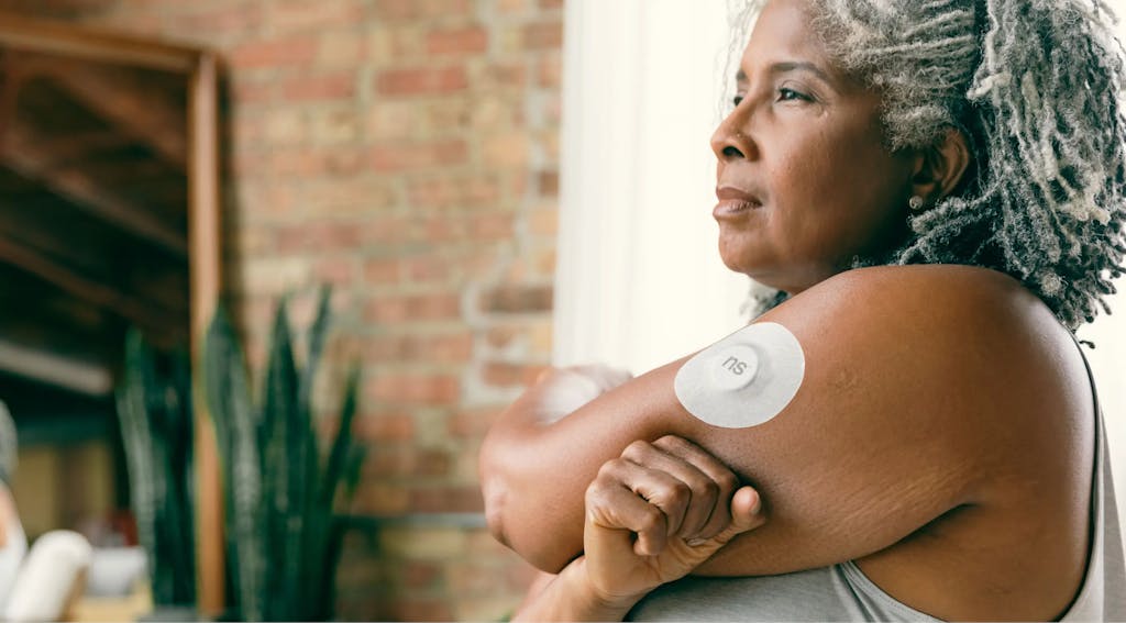 A mature woman with gray hair wearing a continuous glucose monitoring device on her arm.