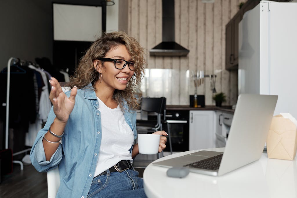 Smiling woman with glasses having a video call from home, enjoying a cup of coffee in her kitchen.